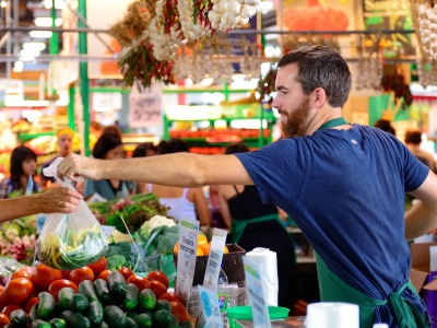 A vendor at a farmer's market hands a bag of vegetables to a customer