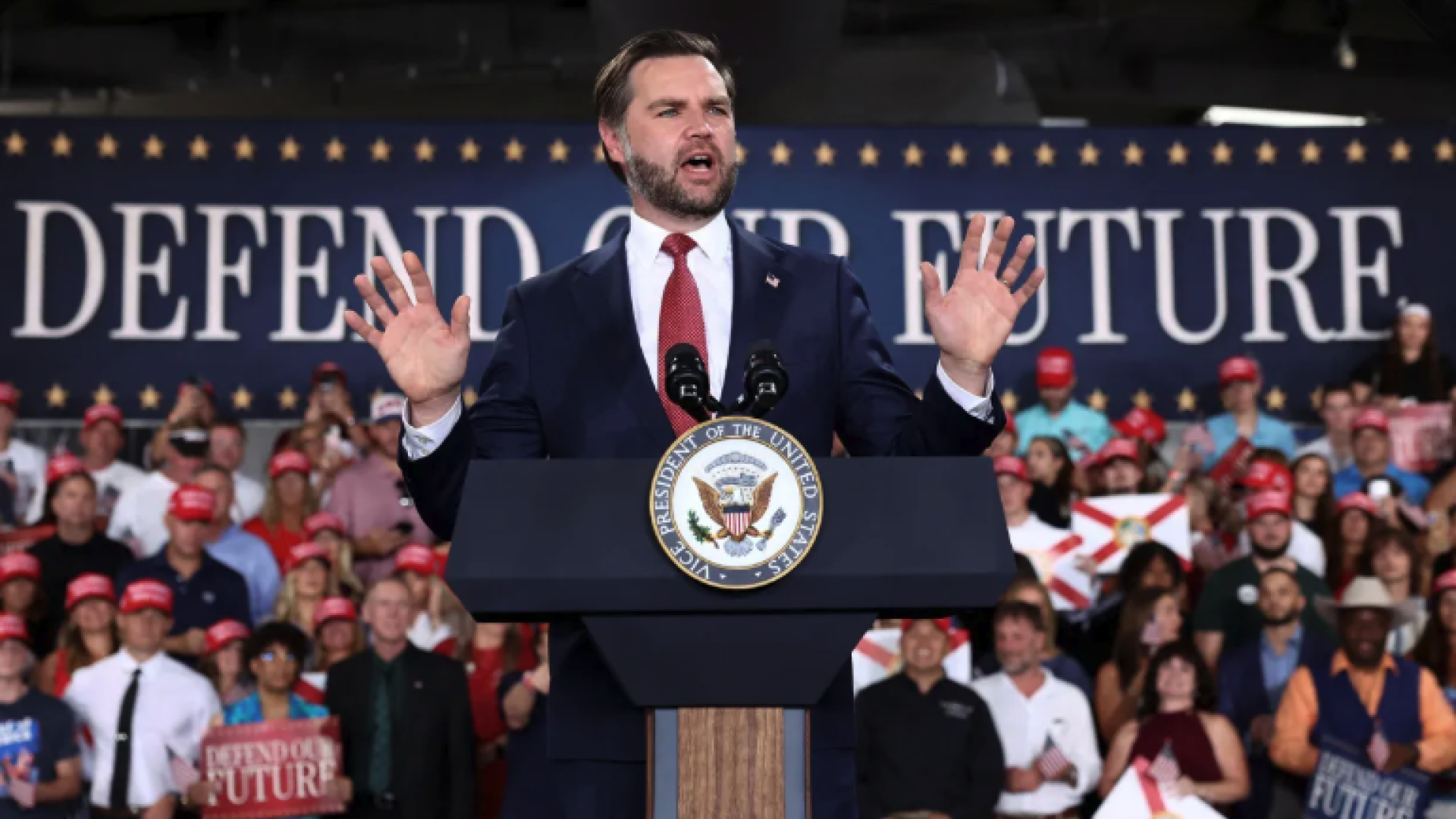 JD Vance speaks at a rally in front of a large crowd and a banner that reads "Defend the Future"