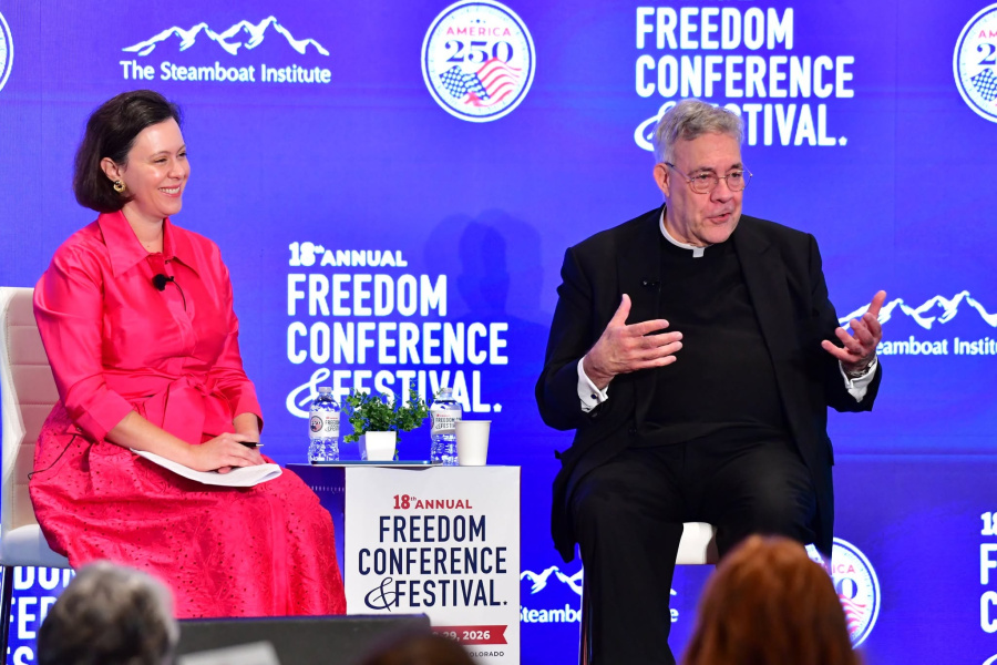 Father Sirico speaks on stage to a crowd in front of a blue LED screen with conference logos